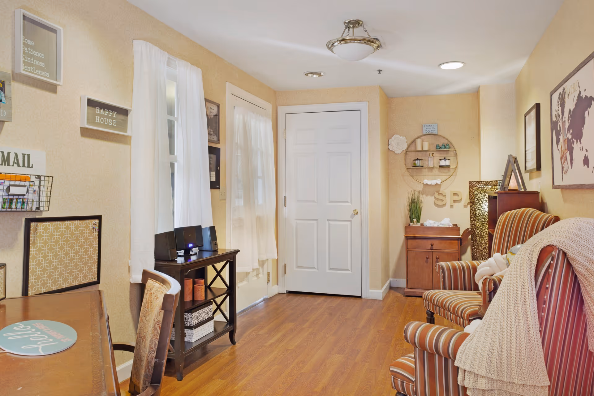 A cozy interior room with beige walls and wooden flooring. The room features a striped armchair with a knitted blanket draped over it, a small wooden cabinet with decorative items, and a wall-mounted circular shelf with spa-related items. There is a white door in the center background, a window with white curtains, and a small table with a chair on the left side. Various framed signs and decorations are hung on the walls.