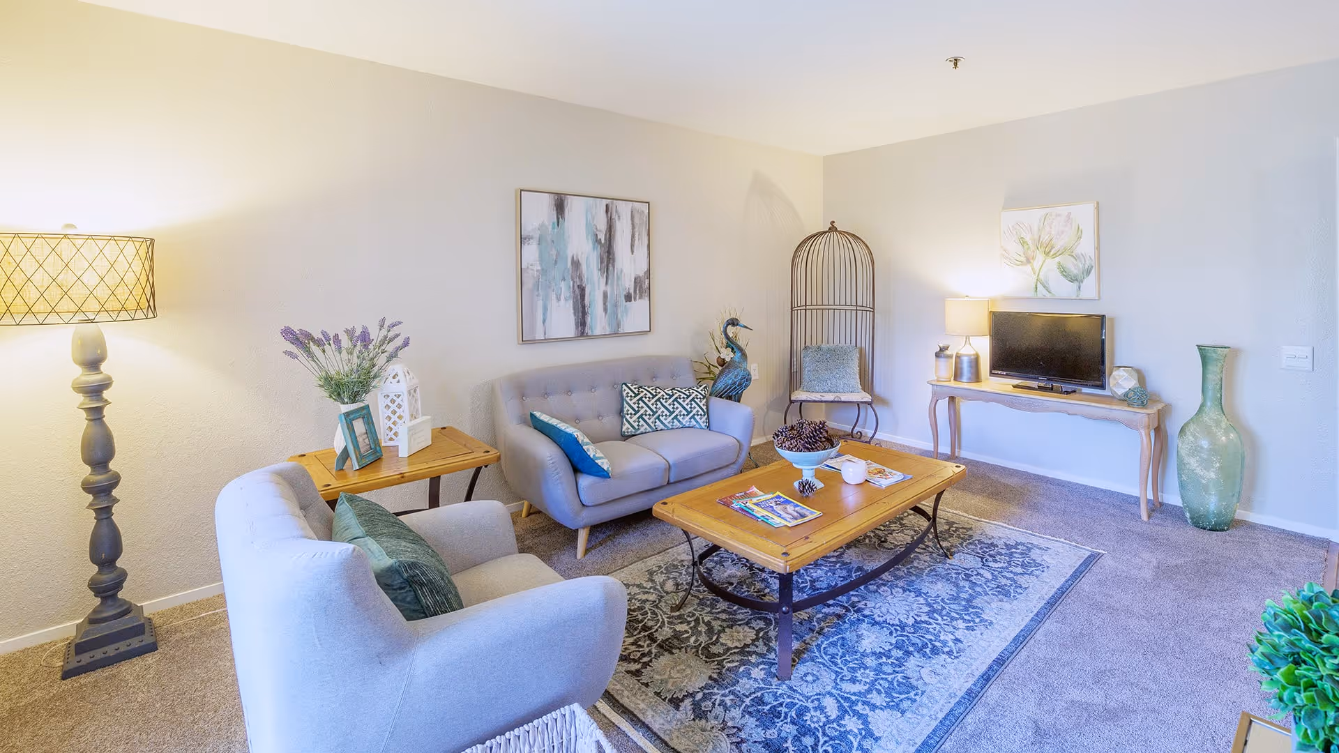A cozy living room with a light gray armchair and loveseat, both adorned with decorative pillows. A wooden coffee table with magazines and a bowl of pinecones sits on a patterned rug. The room features a tall floor lamp, a side table with a vase of flowers and picture frames, a TV on a wooden console table, a large green vase, and wall art. The walls are light-colored and the floor is carpeted.
