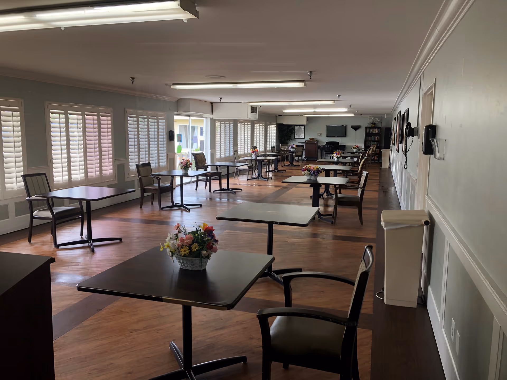 A spacious dining room with multiple square tables and chairs arranged neatly. Each table has a small floral centerpiece. The room has large windows with white shutters allowing natural light to enter. The floor is wooden, and the walls are light-colored with some framed pictures. There are fluorescent lights on the ceiling and a television mounted on the far wall.