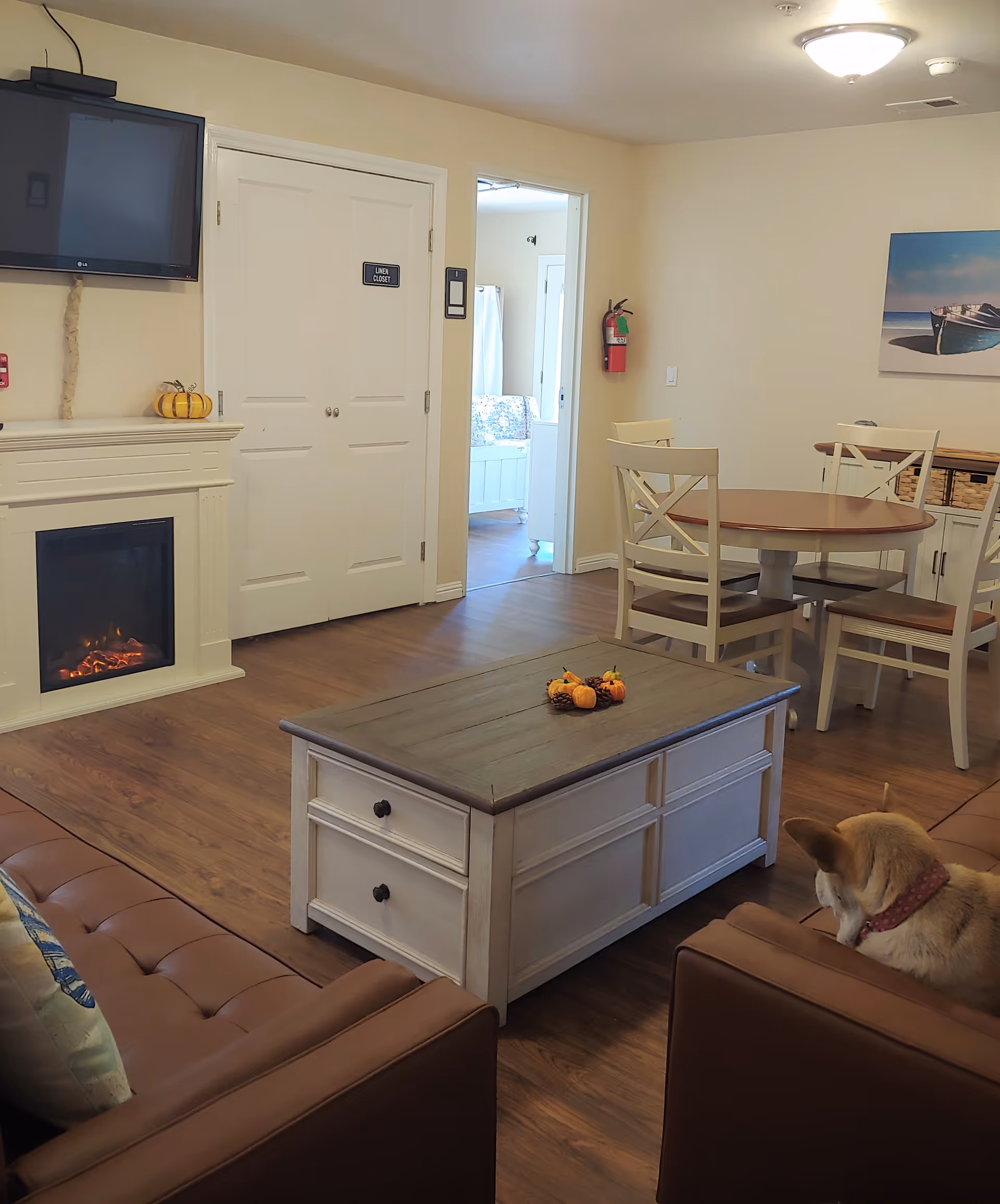 Cozy living room and dining area with a fireplace and wall-mounted TV, a wooden coffee table, round dining table, and a dog on a couch.