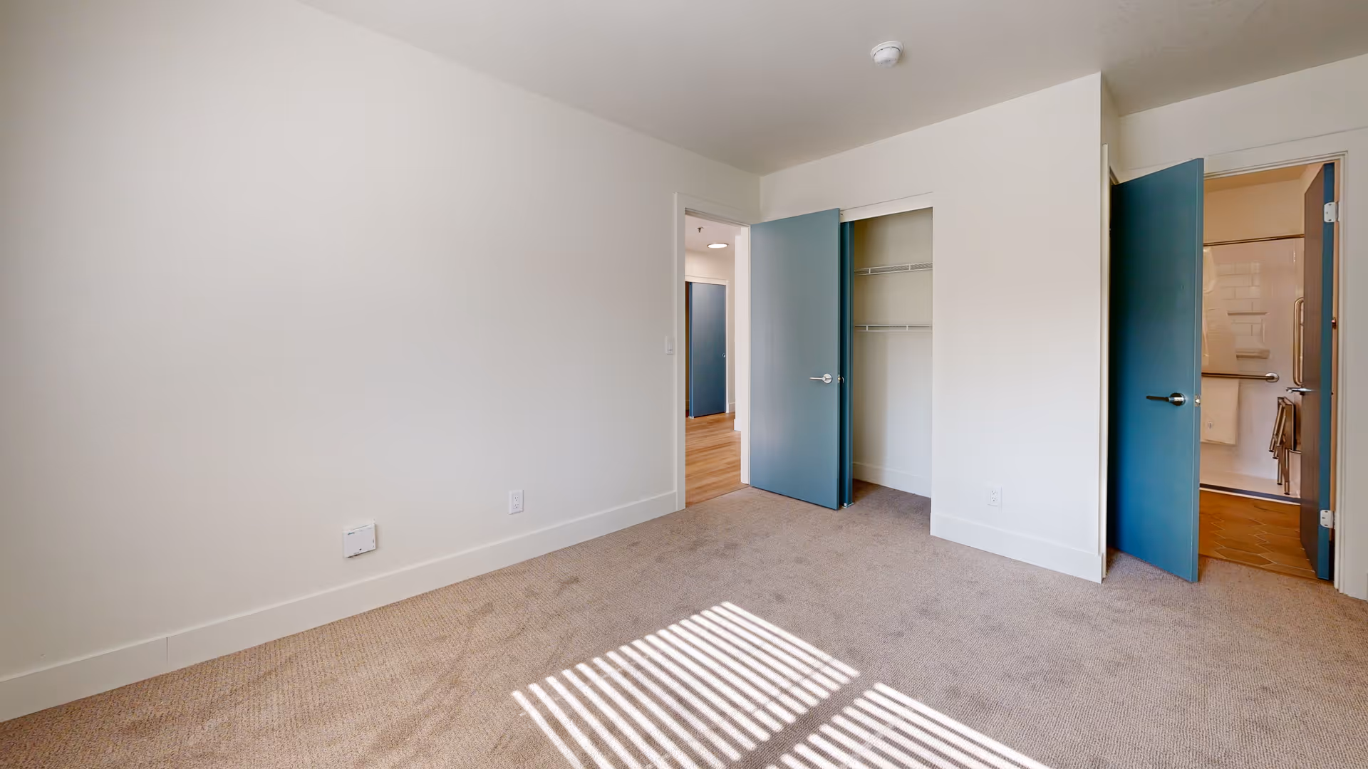 Empty bedroom with beige carpet, white walls, and two blue doors. One door leads to a closet with wire shelving, and the other door opens to a bathroom with a tiled floor and shower. Sunlight casts shadows of window blinds on the carpet.