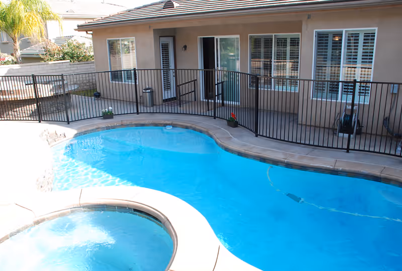 Outdoor swimming pool and hot tub area enclosed by a black safety fence, adjacent to a beige building with windows and a door, with some plants and a palm tree visible in the background.