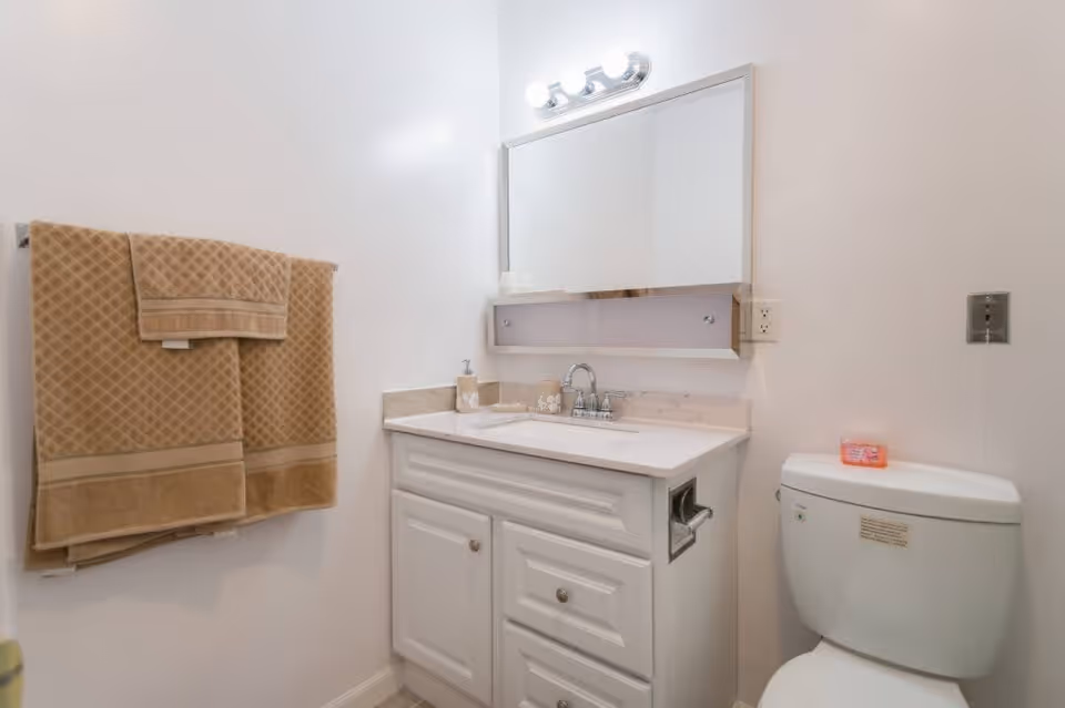 A clean bathroom featuring a white vanity with a marble countertop, a sink with a faucet, a large mirror above the sink with three light bulbs on top, a toilet with a pink soap bar on the tank, and beige towels hanging on a towel rack on the wall.