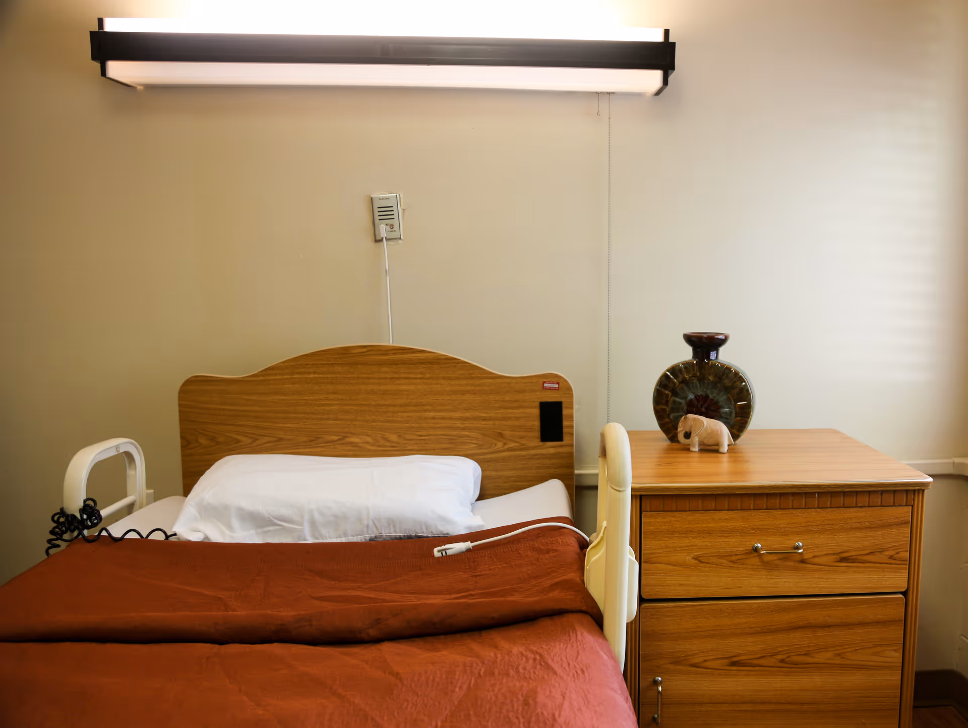 A single bed with a wooden headboard, white pillow, and brown blanket in a room. Next to the bed is a wooden nightstand with a decorative vase and a small elephant figurine on top. A wall-mounted light fixture is above the bed.