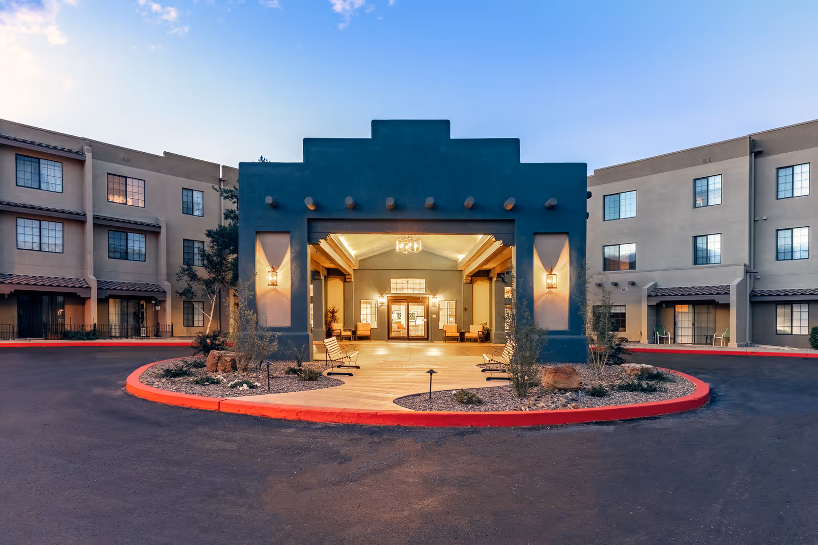 Entrance of a senior living facility named Fairwinds - Rio Rancho, featuring a covered drop-off area with benches, decorative lighting, and a circular driveway bordered by red curbs. The building has multiple windows and a southwestern architectural style.