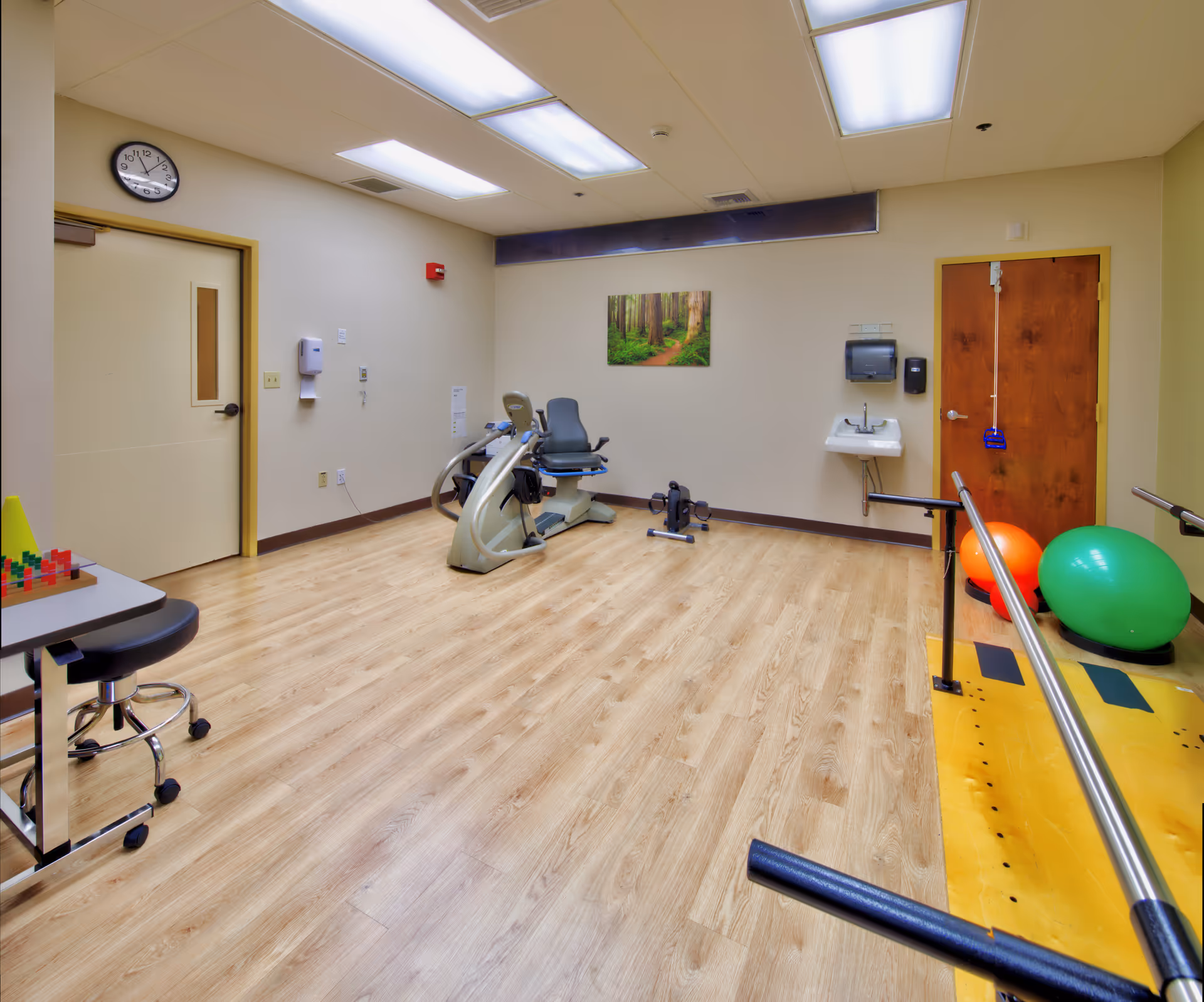 A rehabilitation room with wooden flooring, exercise equipment including a stationary bike, parallel bars for walking support, large exercise balls, a small sink with soap and paper towel dispensers, a clock on the wall, and a nature-themed picture hanging on the wall.