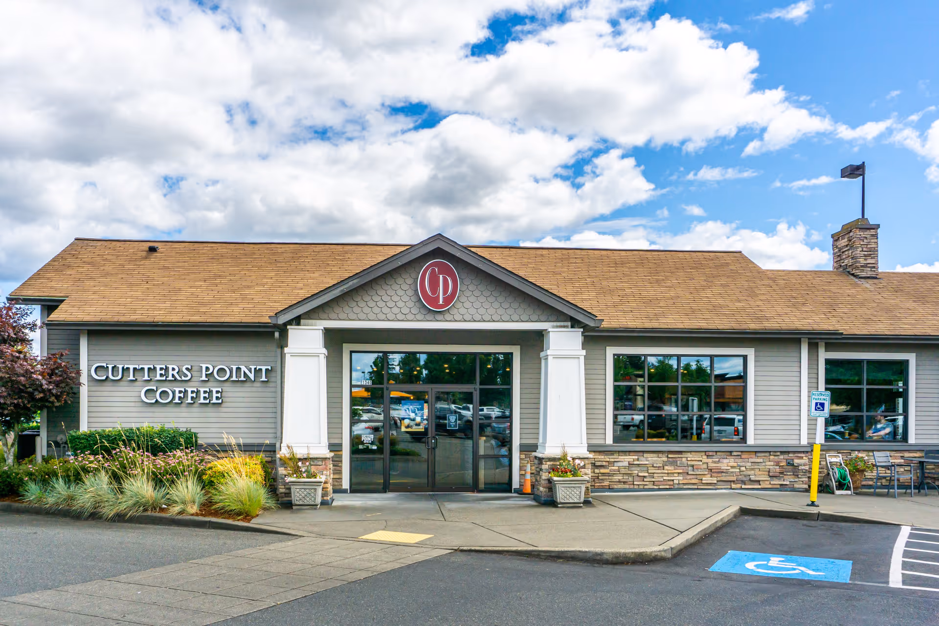 Exterior view of Cutters Point Coffee building with a brown shingled roof, gray siding, stone accents, and large windows. There is a handicapped parking space and a reserved parking sign in front of the building. The sky is partly cloudy with patches of blue.