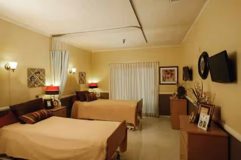 Two beds in a beige convalescent bedroom with lamps, curtains, and dressers.