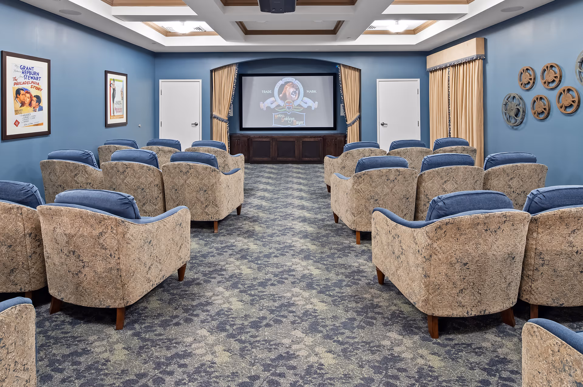 A cozy movie theater room with multiple rows of cushioned armchairs facing a large screen displaying the MGM logo. The walls are painted blue and decorated with vintage movie posters and film reel wall art. The ceiling has recessed lighting with wooden accents, and the floor is carpeted with a patterned design.