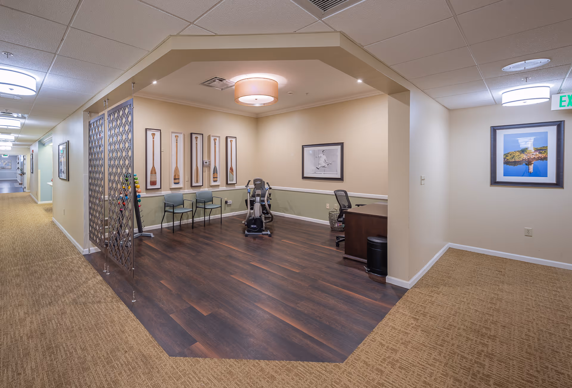 Interior view of a senior living facility hallway leading to a small exercise area with two chairs, an exercise bike, and framed artwork of paddles on the wall. The area has dark wood flooring and is separated from the carpeted hallway by a decorative metal screen. There is a desk with an office chair and a framed picture on the adjacent wall.