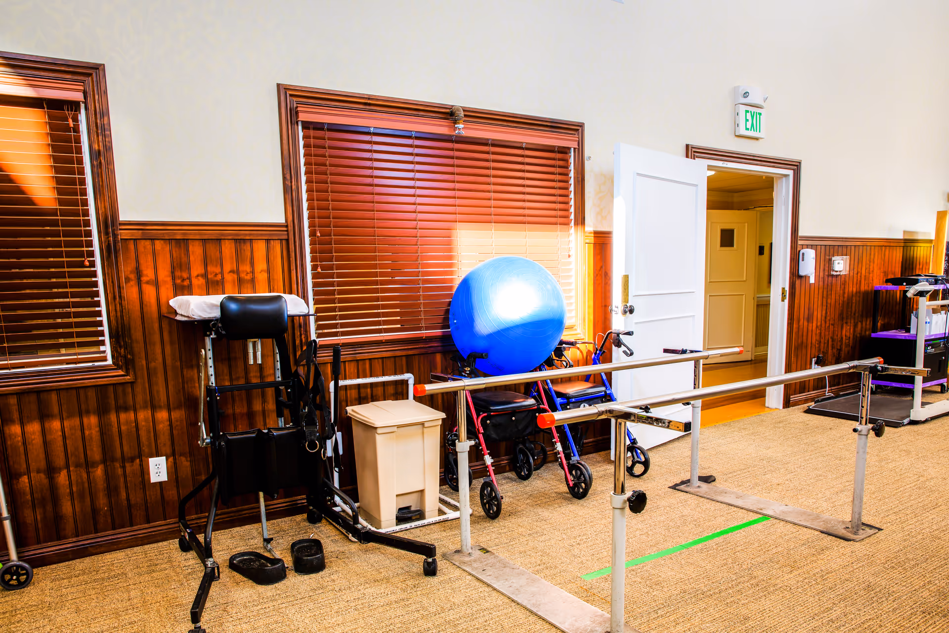 A rehabilitation or physical therapy room with wooden panel walls and two windows with closed blinds. The room contains parallel bars for walking exercises, a large blue exercise ball, a walker, a standing frame, and a beige trash bin. There is an open white door leading to another room, and an exit sign above the door.