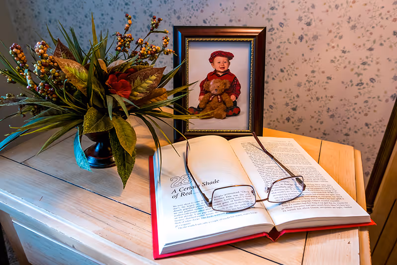 A wooden table with an open book titled 'A Certain Shade of Red' with reading glasses resting on it. Behind the book is a framed photo of a smiling baby wearing a red outfit and holding a teddy bear. To the left of the photo is a decorative vase with artificial green leaves and berries. The background shows a wall with floral wallpaper.