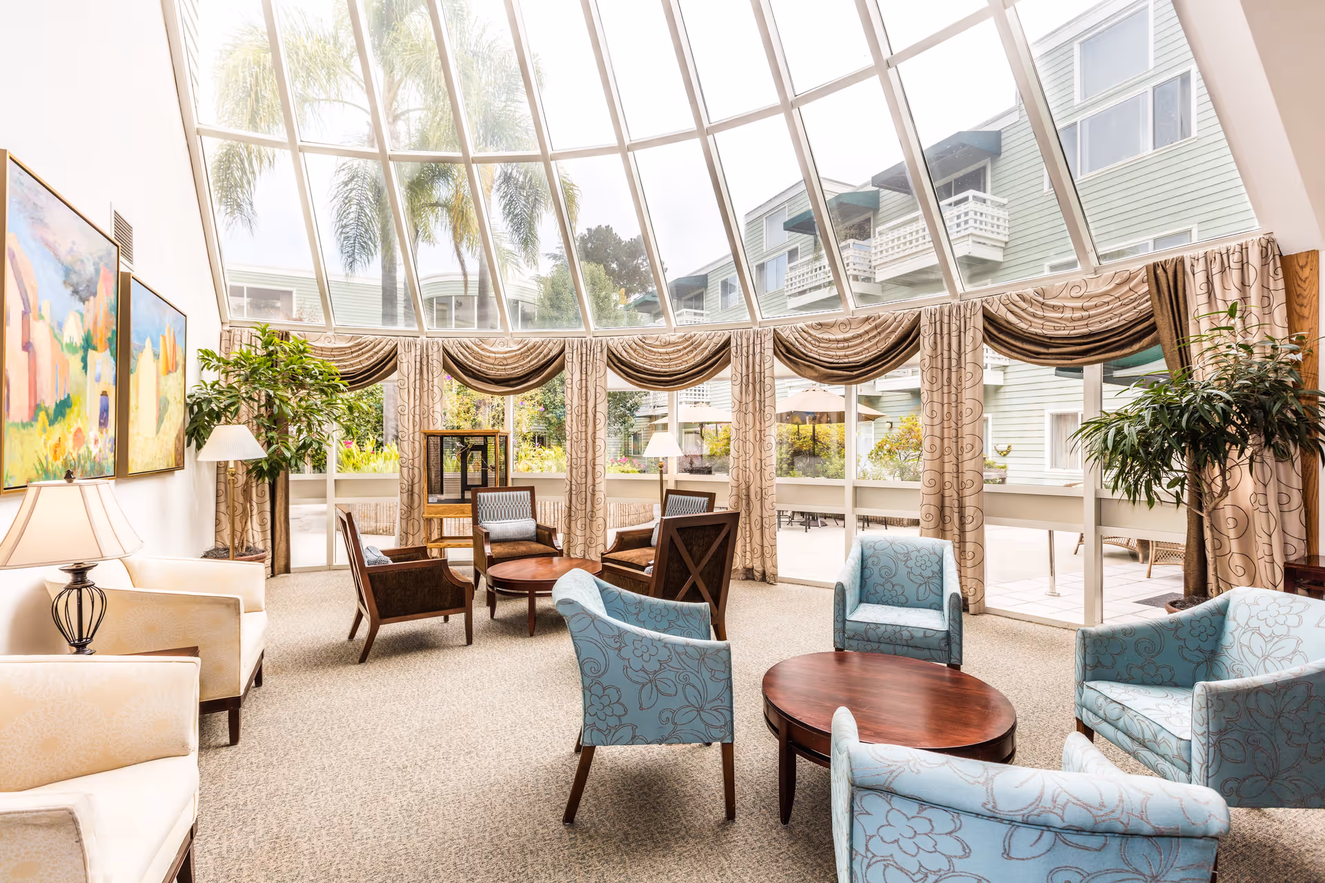 Bright and spacious living room with large floor-to-ceiling windows and a glass ceiling, allowing natural light to fill the space. The room is furnished with multiple armchairs and small round wooden tables arranged for conversation. There are patterned curtains, indoor plants, and colorful paintings on the walls. Outside the windows, a patio area and neighboring building are visible.