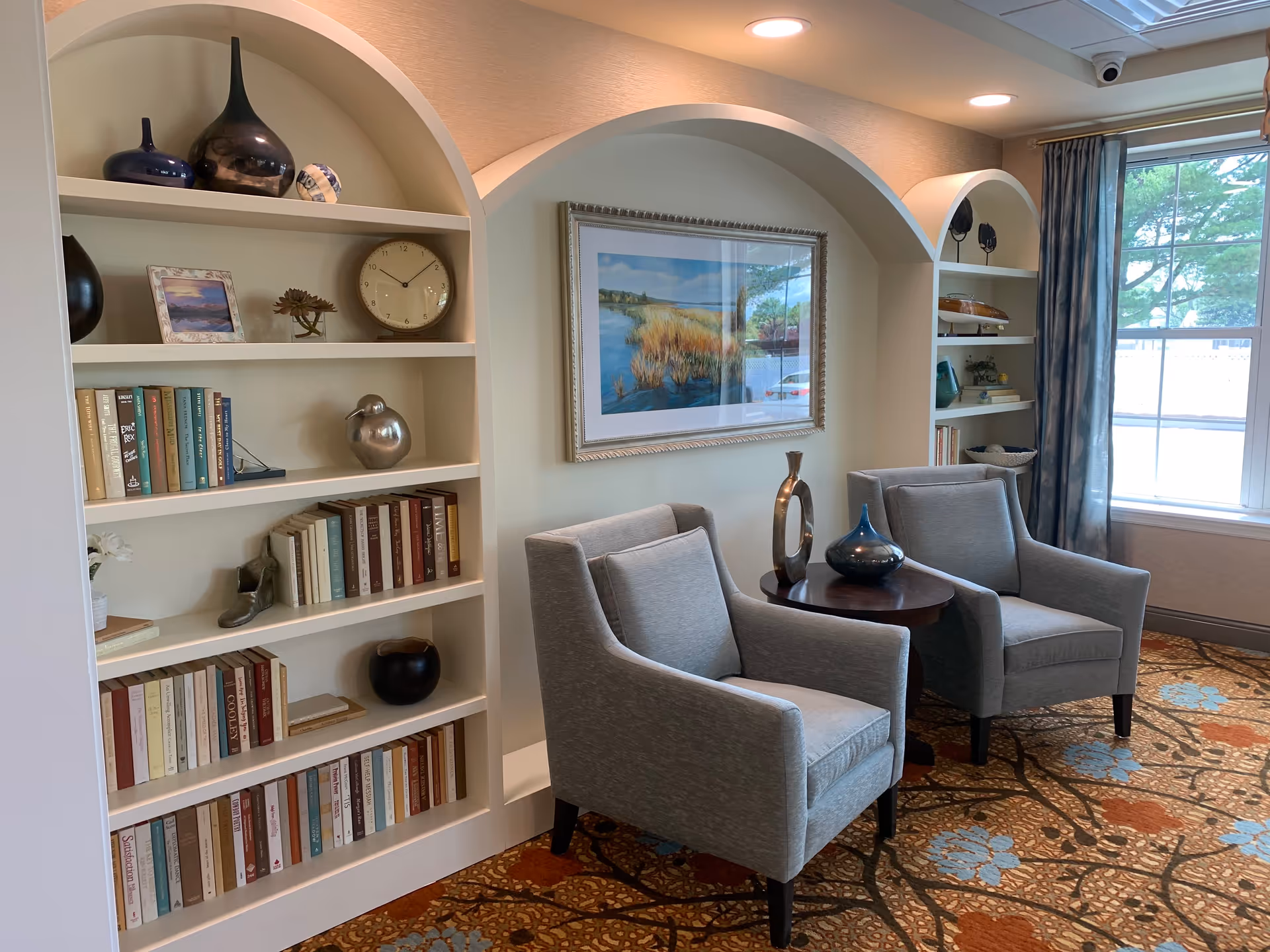 A cozy sitting area with two gray armchairs placed around a small round wooden table holding decorative vases. Behind the chairs are built-in white shelves with various books, decorative objects, and a clock. A framed landscape painting hangs on the wall between two arched alcoves. A large window with blue curtains lets in natural light, and the floor is covered with a patterned carpet featuring blue and orange floral designs.
