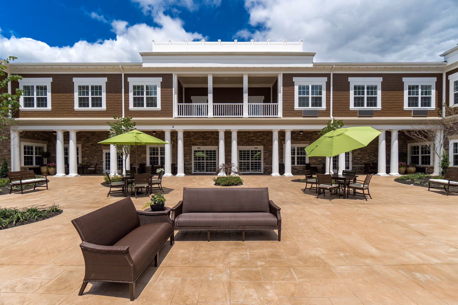 Sunlit courtyard with wicker seating, green umbrellas, and a two-story assisted living building with columns and white-trimmed windows.