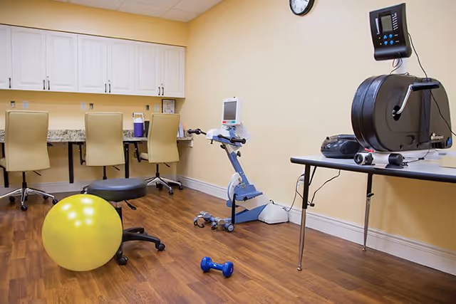 A room with wooden flooring featuring exercise equipment including a yellow exercise ball, a blue dumbbell, a stationary exercise bike, and a rowing machine on a table. There are three beige office chairs in front of a counter with white cabinets above. A clock is mounted on the wall.