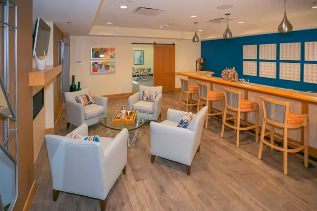 A modern communal lounge with four armchairs around a glass coffee table and a wooden bar with stools against a teal accent wall.