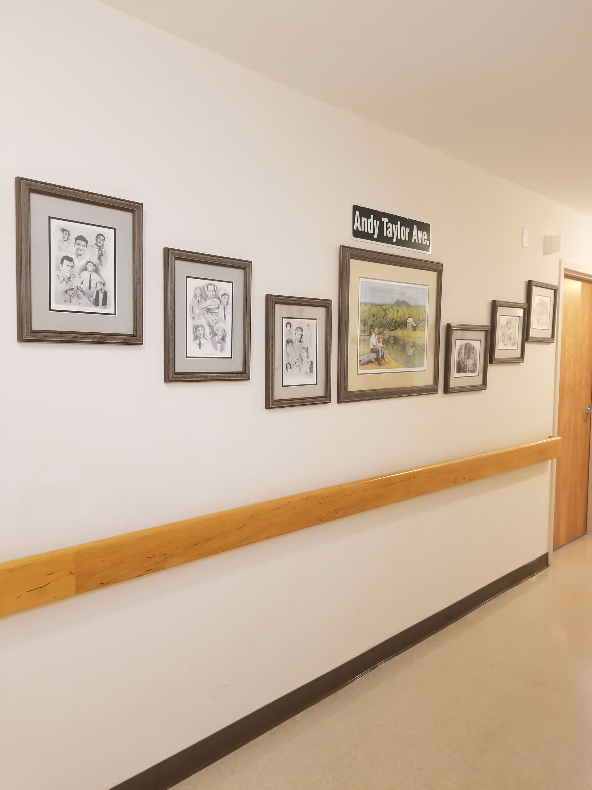 A hallway with a wooden handrail along the wall and several framed pictures hanging above it. One of the frames contains a sign that reads 'Andy Taylor Ave.' The hallway has a light-colored floor and walls, with a closed wooden door at the end.