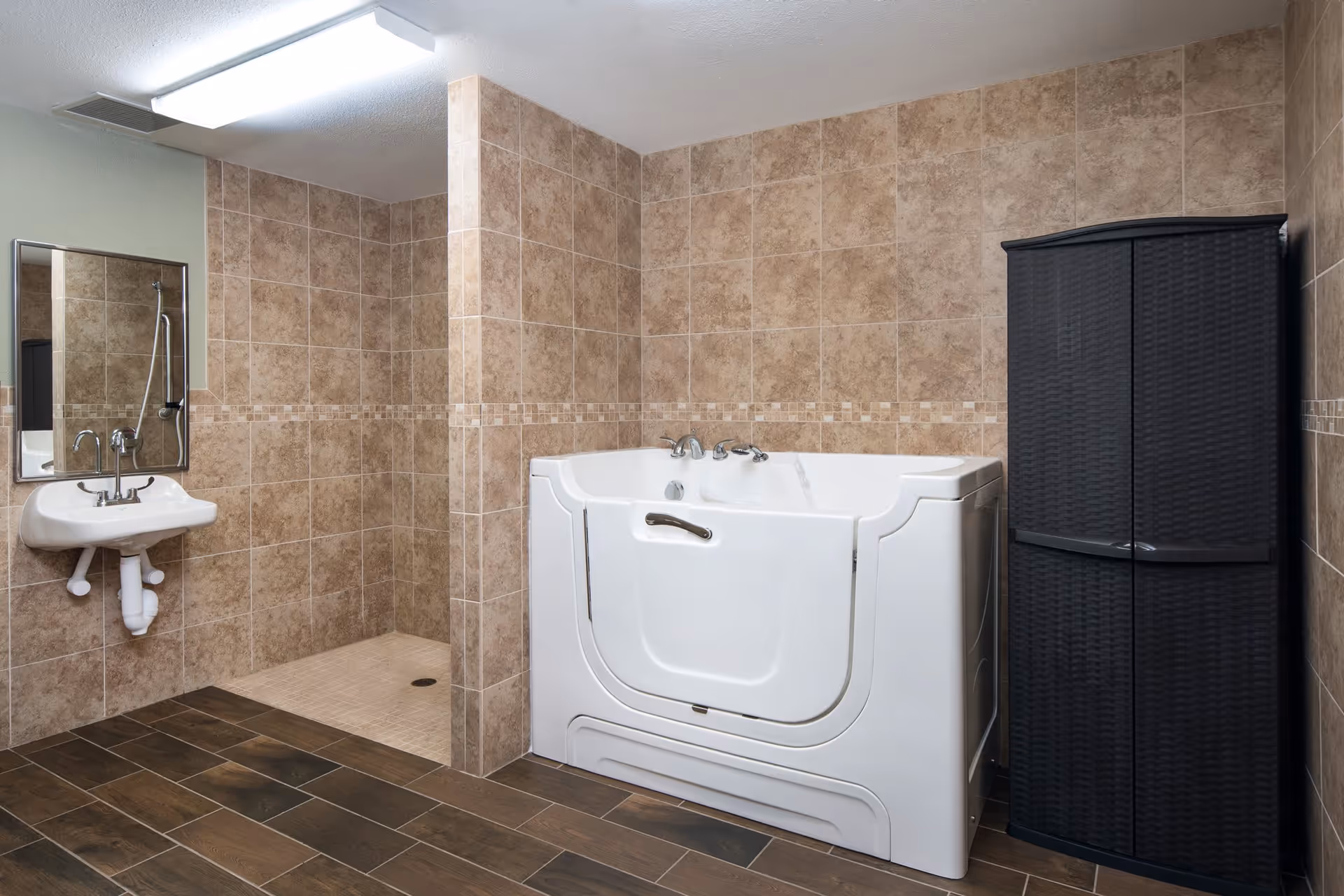 A bathroom with beige tiled walls and dark wood-like tiled floor featuring a walk-in shower area with no door, a white walk-in bathtub with a door and handrails, a small white wall-mounted sink with a mirror above it, and a tall black storage cabinet.