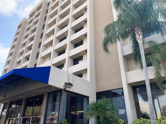 Exterior view of a multi-story beige building with balconies, a blue awning over the entrance, and palm trees in front under a partly cloudy sky.