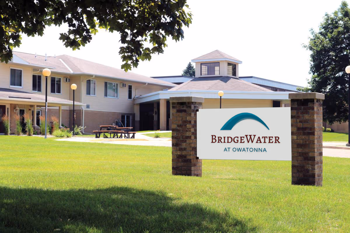 Exterior view of the BridgeWater at Owatonna senior living facility showing a two-story building with beige siding and brick accents, surrounded by green grass and trees, with a sign in the foreground displaying the facility's name.