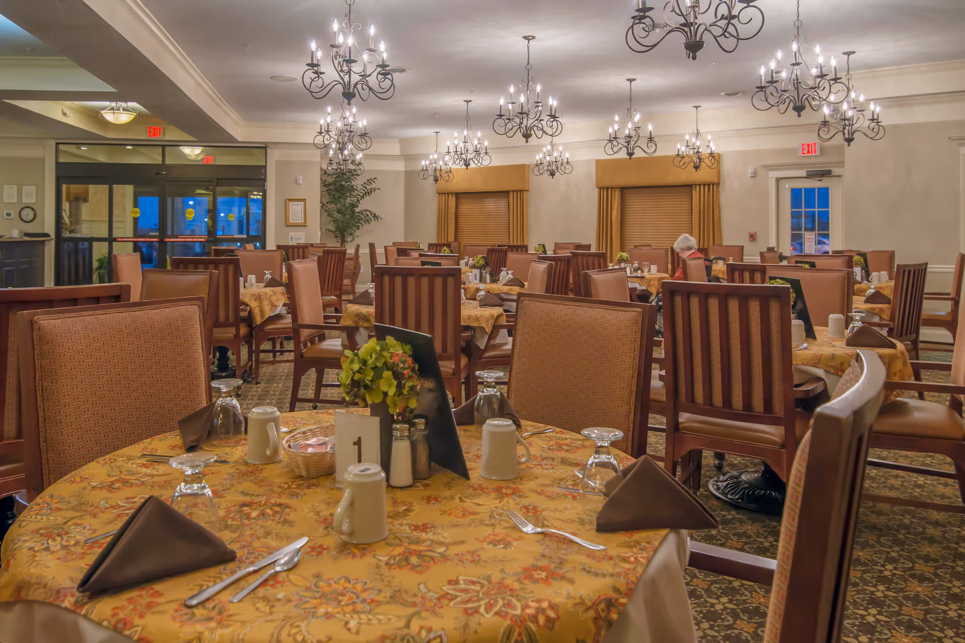 A spacious dining room in a senior living facility with multiple tables covered in floral tablecloths, each set with utensils, glasses, napkins, and condiments. The room is warmly lit by several ornate chandeliers hanging from the ceiling. There are wooden chairs with cushioned seats around the tables, and a few windows with closed blinds. One person is seated at a table in the background.