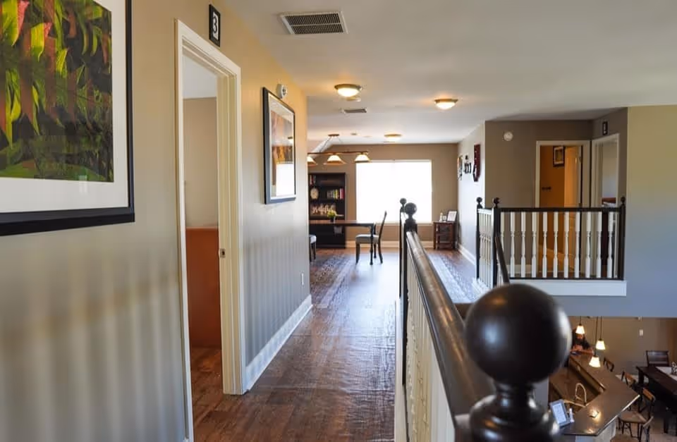 Interior hallway of a senior living facility with wooden flooring, beige walls, framed artwork, and a railing on the right side overlooking a lower level with tables and chairs. There are doors along the hallway and a window at the far end letting in natural light.
