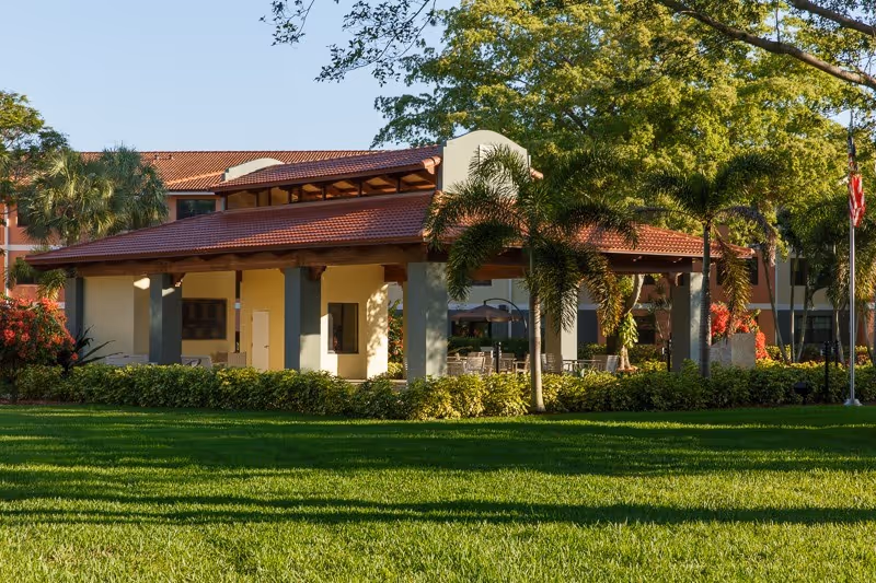 Covered outdoor pavilion and entrance with a red tiled roof, palm trees and a manicured green lawn.