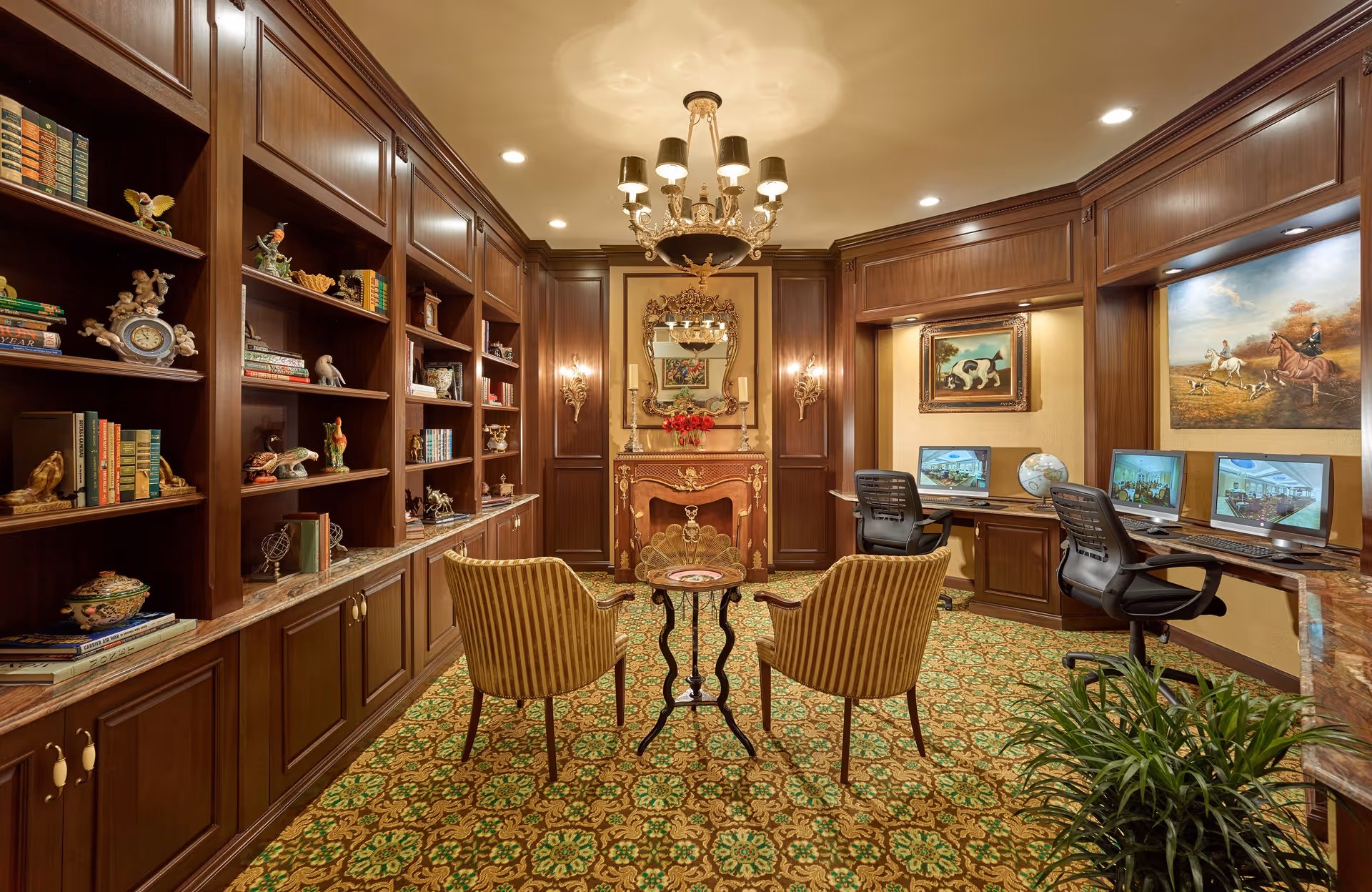 A luxurious interior room with dark wood paneling and built-in bookshelves filled with books and decorative items. Two striped armchairs face a small round table in the center of the room. A chandelier hangs from the ceiling, and there is an ornate fireplace with a decorative mirror above it. On the right side, there are two desks with computers and office chairs, and paintings hang on the walls. The floor is covered with a patterned carpet, and a green plant is visible in the foreground.