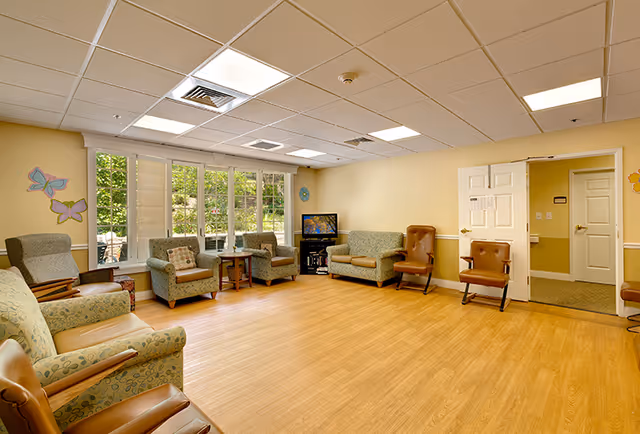 A bright and spacious common living area in a memory care community with multiple armchairs and sofas arranged around the room. Large windows on one wall let in natural light and offer a view of greenery outside. A small TV is placed on a stand in the corner near an open door leading to another room. The walls are painted light yellow and decorated with colorful butterfly wall art.