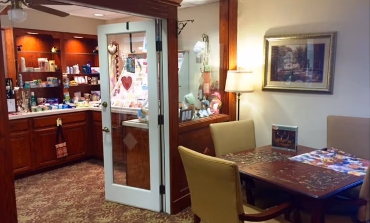 Interior view of a room in Bethany Village showing a small seating area with a wooden table and four beige chairs. The table has a partially completed jigsaw puzzle and a puzzle box on it. To the left, there is a glass door leading to a small shop or pantry area with wooden cabinets and shelves stocked with various items. A floor lamp and a framed picture are on the wall behind the seating area.