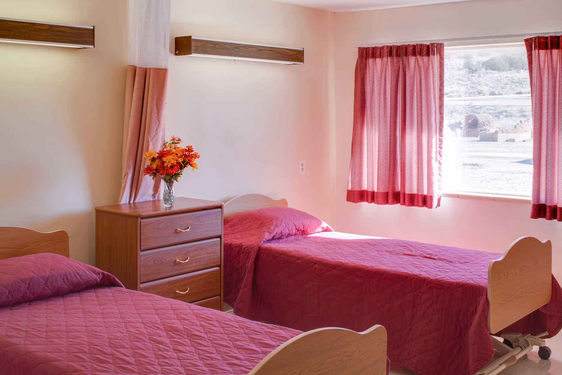 Two single beds with pink bedspreads, a wooden dresser topped with a vase of flowers, and a window with curtains in a nursing facility room.