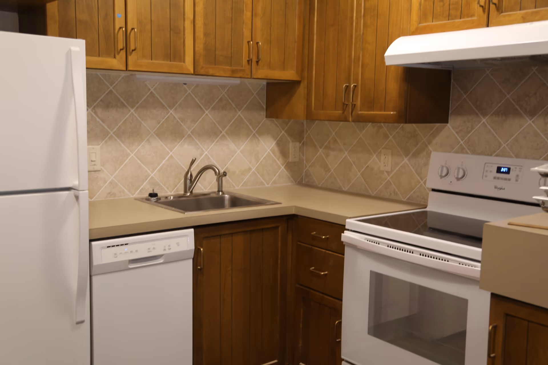 A kitchen with wooden cabinets, a white refrigerator, a white dishwasher, a stainless steel sink with a faucet, a white electric stove with an oven, and beige tiled backsplash.