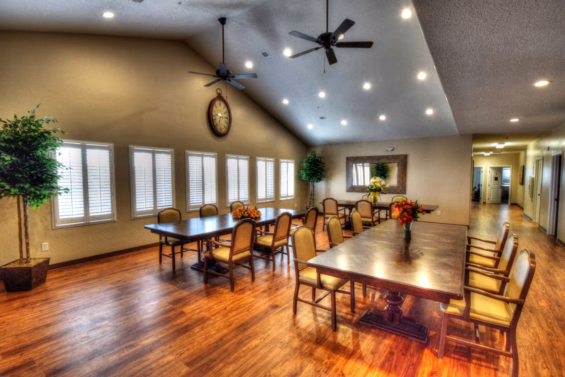 Spacious dining room with long wooden tables and upholstered chairs, wood floors, ceiling fans, and shuttered windows.