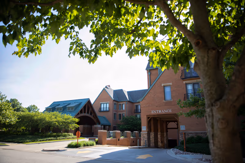 Exterior view of a senior living facility building with a brick facade, multiple windows, and a covered entrance. There is a large tree with green leaves in the foreground and well-maintained landscaping around the building. The word 'ENTRANCE' is visible above the covered driveway.