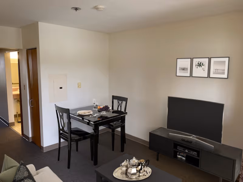 A small living area with a black dining table set for two, a black TV stand with a flat-screen TV, and a coffee table with a silver tea set. There is a beige couch partially visible on the left side. The walls are light-colored with three framed pictures hanging above the TV. A doorway leads to a bathroom area with a sink and mirror visible.