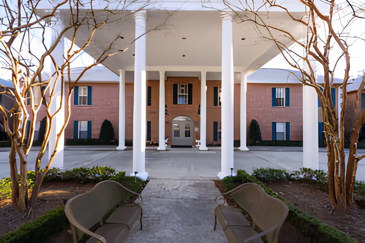 Entrance of a senior living facility with a covered driveway supported by white columns. There are two benches facing each other on either side of the walkway, with landscaped bushes and leafless trees around them. The building in the background is two stories with pinkish walls and blue window shutters.
