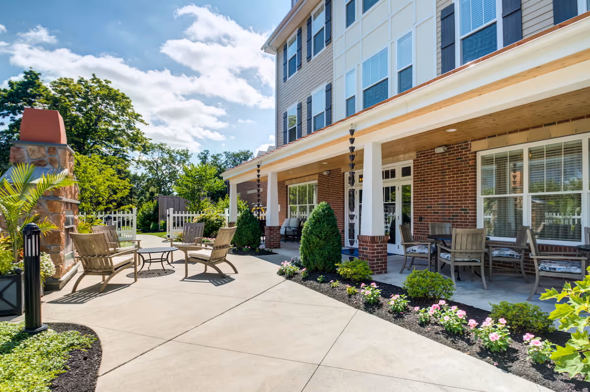 Outdoor patio area of Arbor Terrace Morris Plains featuring a paved walkway, several chairs and tables, a stone fireplace, and well-maintained landscaping with bushes and flowers under a partly cloudy sky.