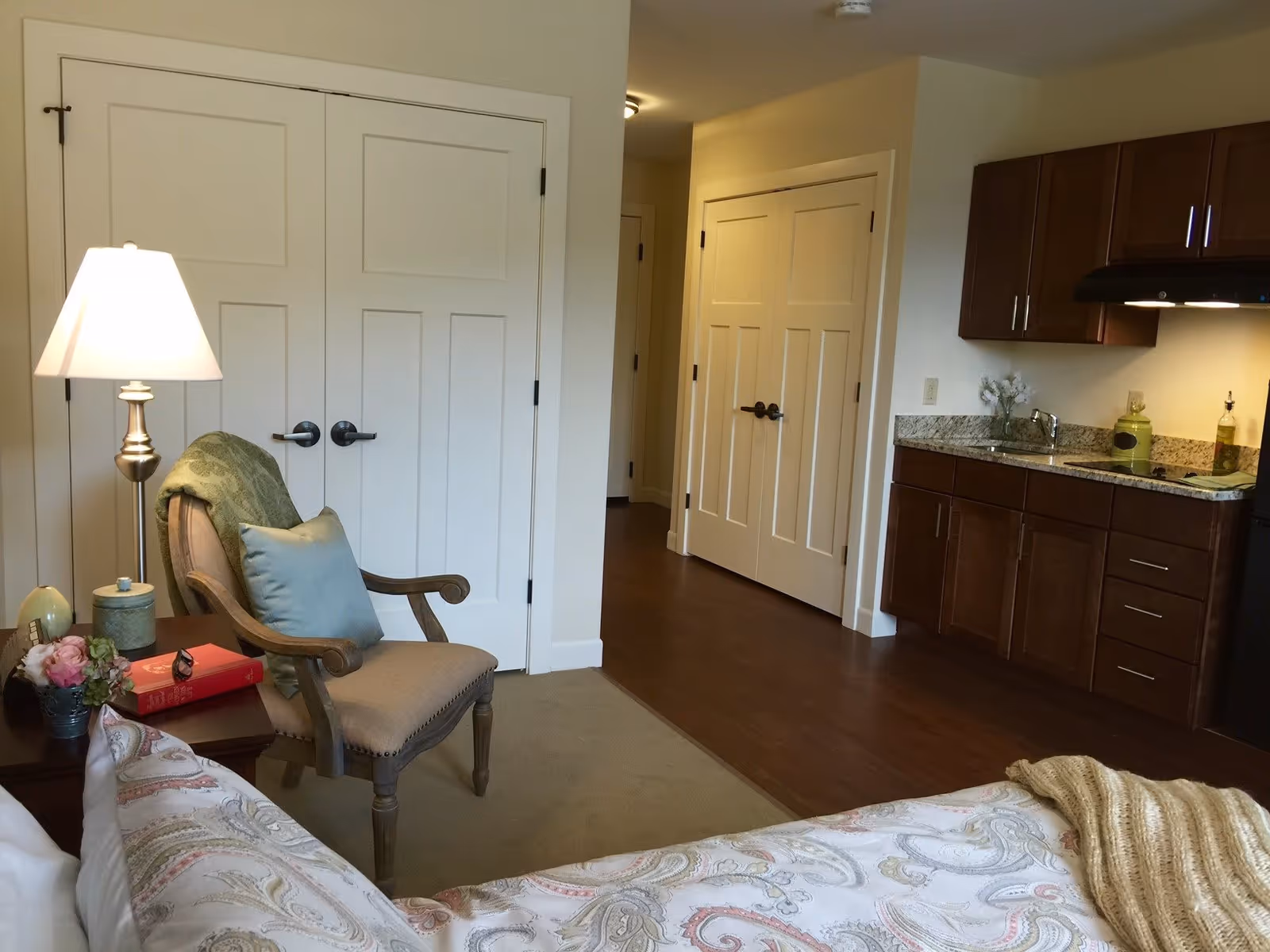 A cozy bedroom area in a senior living facility featuring a bed with patterned bedding, a wooden armchair with a green throw and blue pillow, a side table with a lamp, flowers, and a book. The room has wooden flooring near a kitchenette with dark wood cabinets, a granite countertop, and a sink. There are white paneled double doors and a hallway visible in the background.