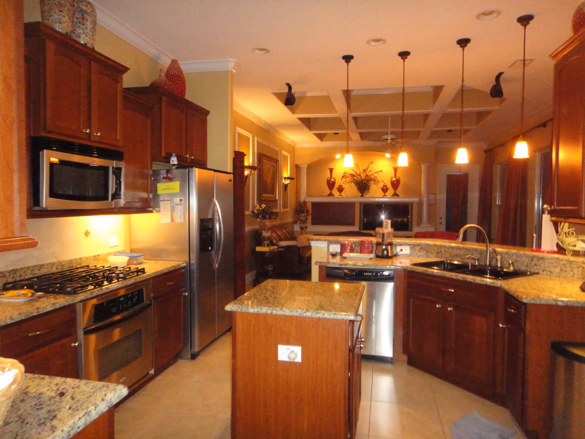 Interior view of a kitchen with wooden cabinets, granite countertops, a stainless steel refrigerator, microwave, oven, and dishwasher. The kitchen opens into a living area with decorative vases and warm lighting from hanging pendant lights and wall sconces.