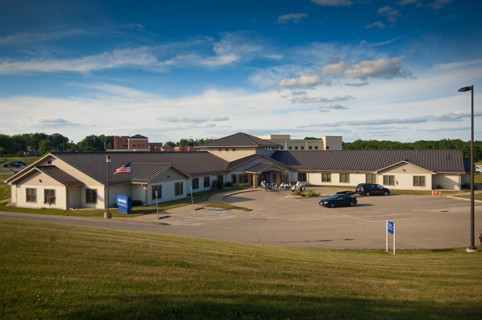 Exterior view of Upland Hills Nursing And Rehab Center, a single-story building with a dark roof and beige walls, surrounded by a parking lot with a few cars and an American flag on a flagpole near the entrance. The sky is partly cloudy with blue patches.