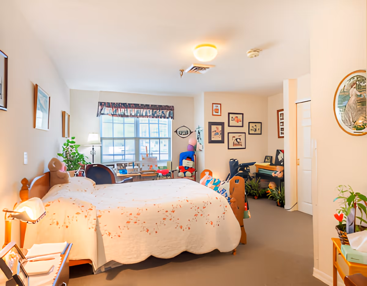 A cozy bedroom with a large bed covered in a white quilt with floral patterns. There is a bedside table with a lamp and some books, a window with a valance letting in natural light, several framed pictures on the walls, a chair with colorful cushions, and various plants and decorative items throughout the room.