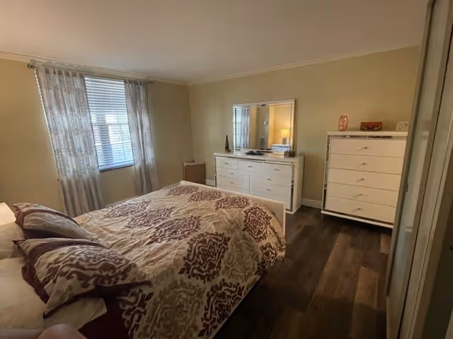 A bedroom with a bed covered in a patterned comforter and pillows, a window with sheer curtains allowing natural light, a white dresser with a mirror, and a tall white chest of drawers on a wooden floor.