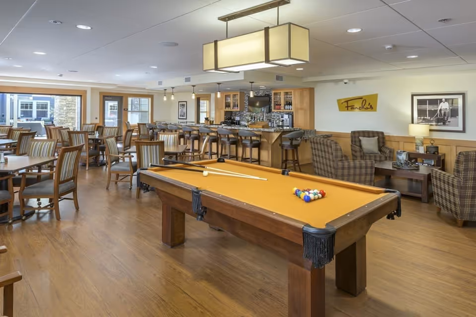Spacious communal lounge with a yellow-felt pool table in the foreground, dining tables, a bar, and cozy seating.