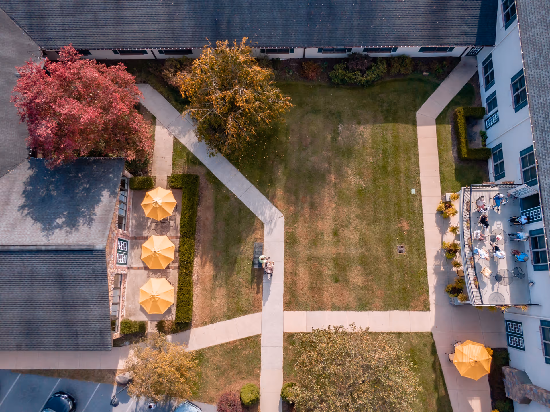 Aerial view of a senior living facility courtyard with green lawns, paved walkways, trees with autumn foliage, and several yellow patio umbrellas. A group of people is gathered on a patio on the right side, and two individuals are sitting on a bench in the center of the courtyard.
