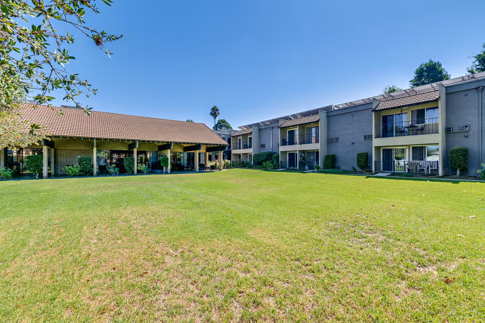 A large green lawn in front of a two-story senior living facility building with balconies and a covered patio area under a clear blue sky.