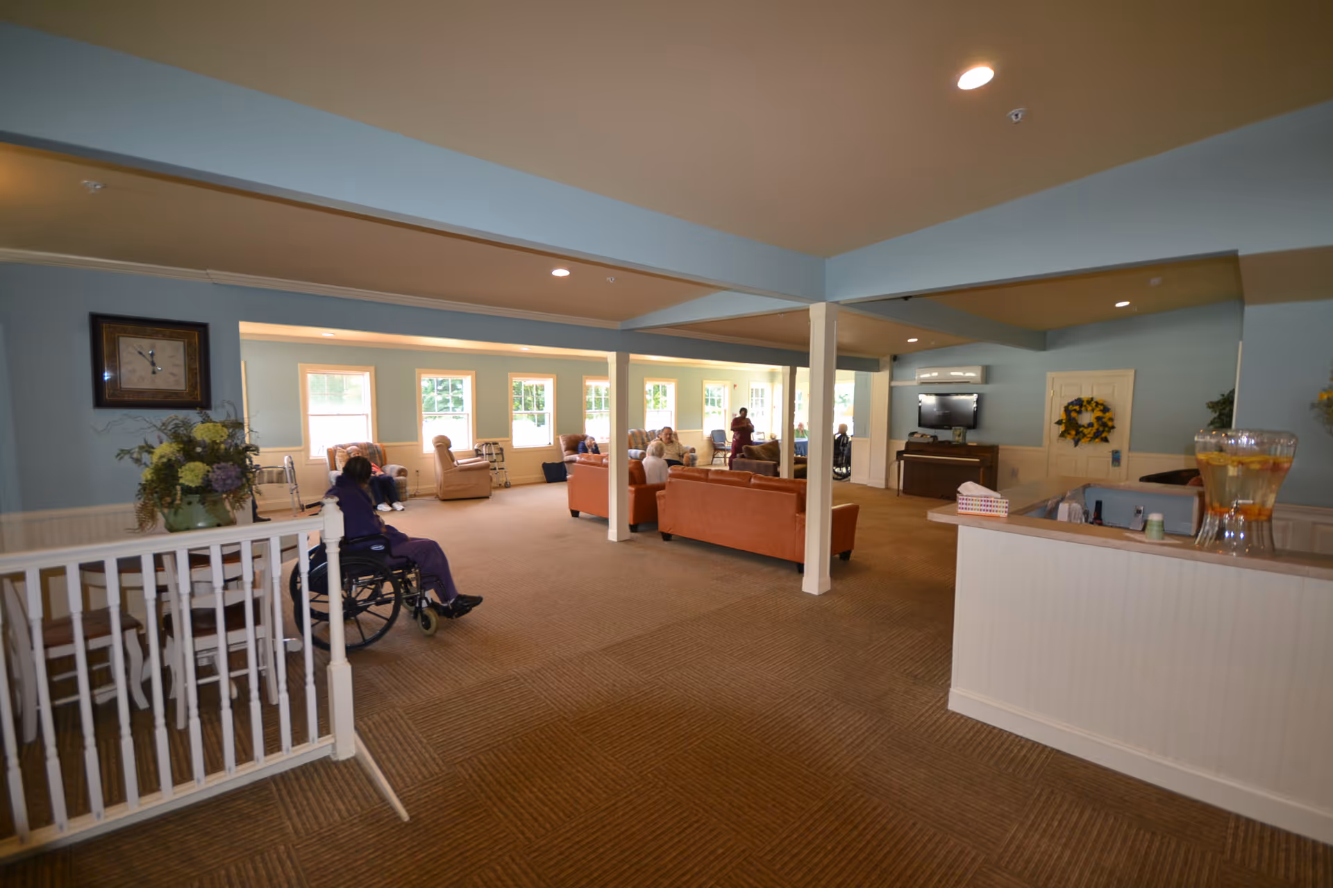 A spacious assisted living common area with light blue walls and beige carpet. Several elderly residents are seated on sofas and armchairs near large windows letting in natural light. A woman in a wheelchair is visible in the foreground. The room has white pillars, a wall-mounted TV, and a counter with a large glass container of infused water.