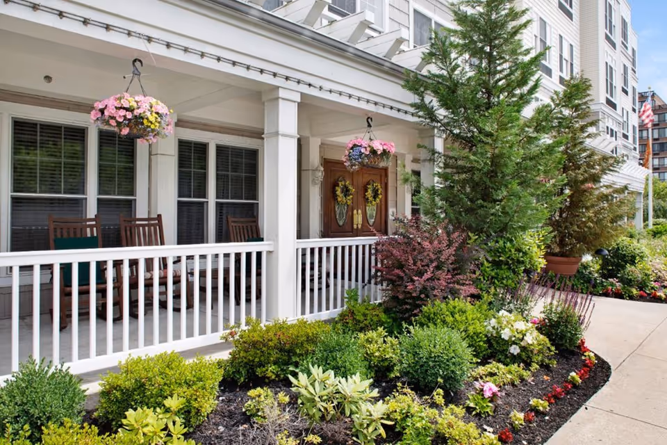 Front porch area of a senior living facility with white railing, wooden rocking chairs, hanging flower baskets, and a variety of green shrubs and flowering plants along the walkway.
