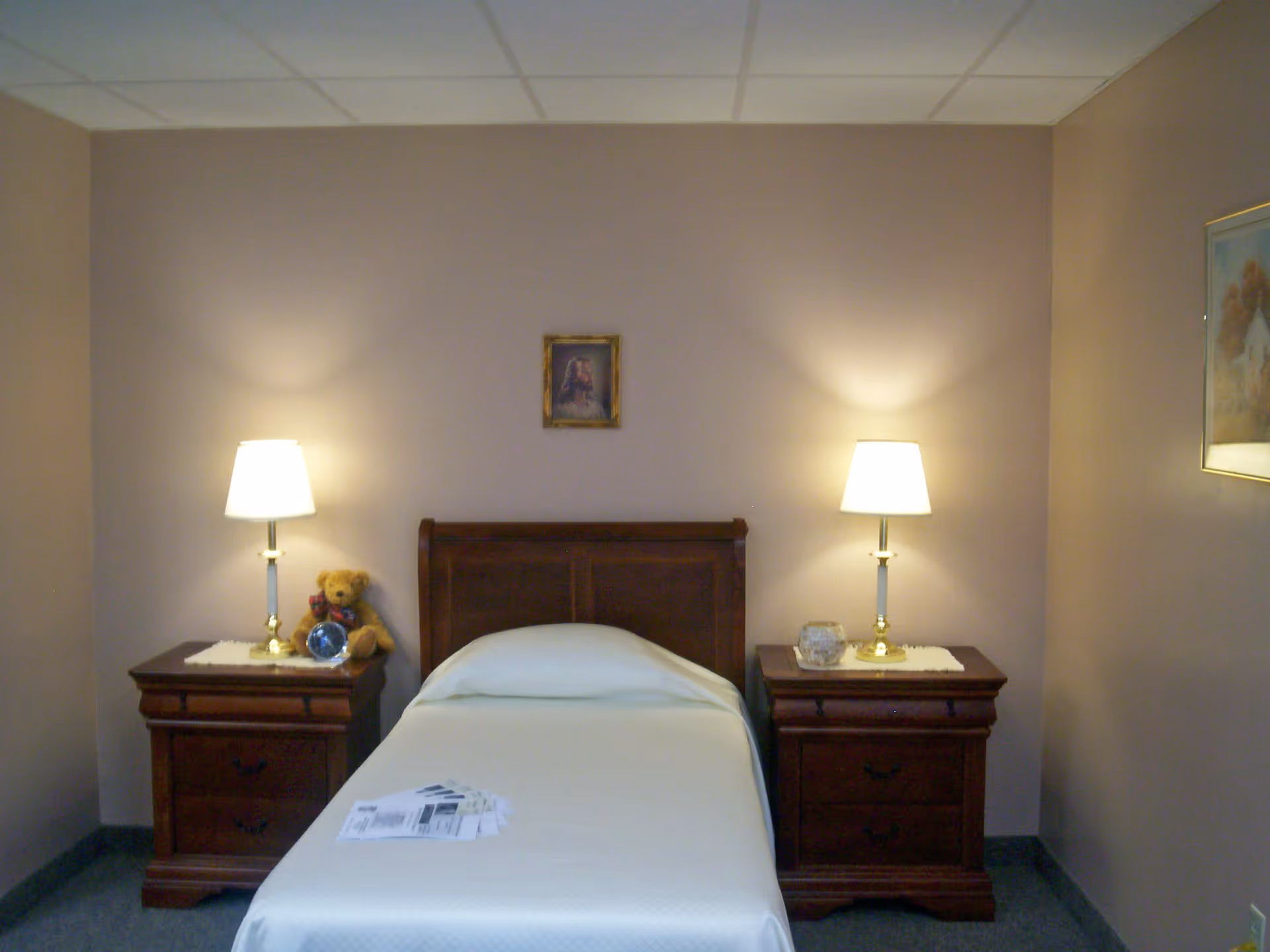 A simple bedroom with a single bed centered between two wooden nightstands. Each nightstand has a lit table lamp. On the left nightstand, there is a teddy bear and an alarm clock. The bed is made with a white bedspread. Above the bed hangs a small framed picture, and there is another framed picture on the right wall. The walls are painted a light beige color.