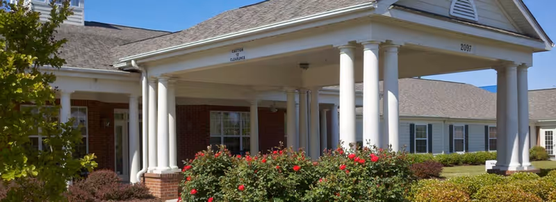 Entrance of a brick building with a white columned portico, windows, and landscaped shrubs and roses under a clear sky.